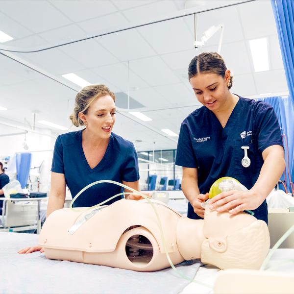 A nursing instructor supervises a student practicing on a medical dummy in a clinical training environment.