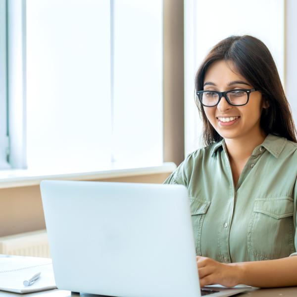 The image shows a woman sitting at a desk, working on a laptop. She is smiling, appearing engaged and focused on the screen. The woman is wearing glasses and a light green shirt, giving her a casual yet professional look.