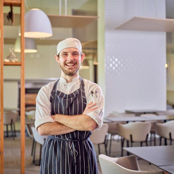 This image showcases a chef standing confidently in a modern dining area. The chef is dressed in a professional uniform, including a striped apron and a white hat. The setting is bright and stylish, with contemporary furniture and lighting, creating a welcoming atmosphere.