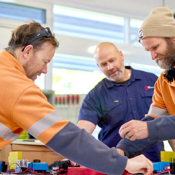 The scene captures three individuals engaging in a hands-on workshop. Two men in the foreground, dressed in casual work attire, are focused on assembling or adjusting components on a table. They are guided by a man in the background, possibly an instructor or supervisor. The room is well-lit with natural light coming through large windows, creating a collaborative and educational atmosphere.