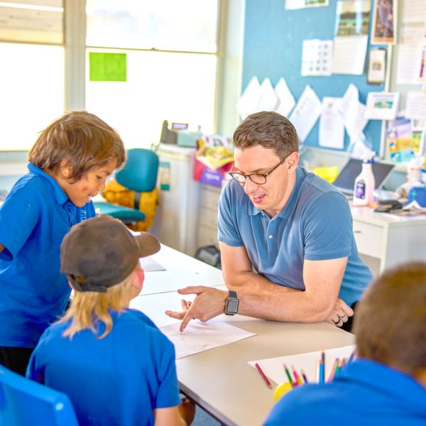 A teacher engages with young students, offering guidance at a classroom table.