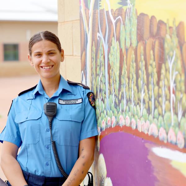 The image shows a person in a blue uniform with a patch that reads "CORRECTIVE SERVICES." The individual is standing outdoors, leaning against a wall with a colorful mural that depicts a landscape scene with trees and a purple area, possibly representing a river or pathway.