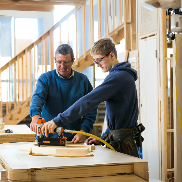 The image shows two individuals in a workshop setting, engaging in a woodworking activity. One person, likely an instructor or mentor, is guiding the other person, who appears to be a student or trainee. The instructor is wearing glasses and a blue sweatshirt, while the trainee is dressed in a hoodie with a tool belt around their waist and protective glasses.